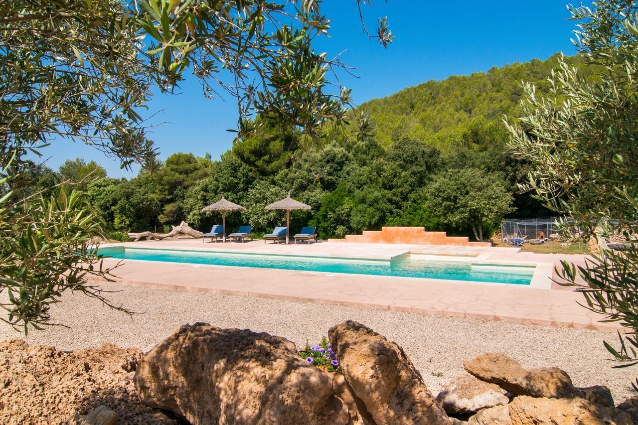 Familienfreundliche Finca auf Mallorca mit großem Pool, Sonnenliegen und Strohdach-Schirmen inmitten mediterraner Natur.